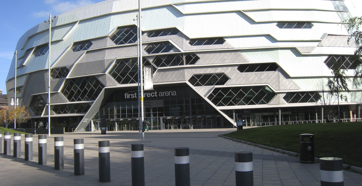 Exterior view of First Direct Arena in Leeds, featuring its unique honeycomb-style geometric façade with angular windows and metallic panels, set under a clear blue sky with surrounding bollards and pedestrian space.