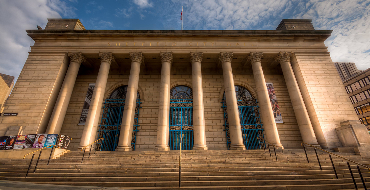 Front view of Sheffield City Hall, a neoclassical building with large stone columns, blue iron gates, and wide steps leading to the entrance. The sky above is partly cloudy with warm evening light casting shadows on the façade.