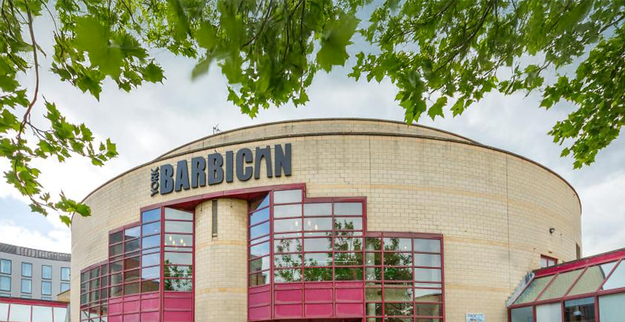 Front view of York Barbican, a curved brick building with large red-framed windows, surrounded by green leafy trees. The venue's name is prominently displayed at the top in bold black lettering.